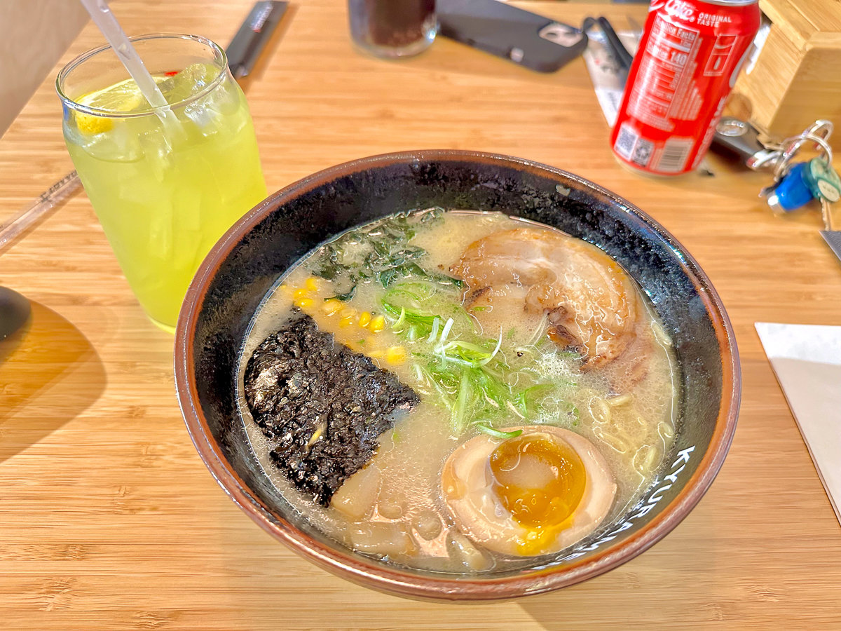 bowl of ramen with toppings on a brown table with a yellow drink